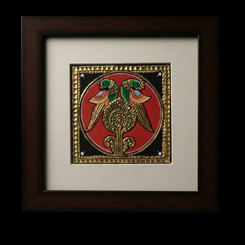 Hand-painted Tanjore painting of two green parrots perched on a golden pedestal within a circular frame, featuring 22K gold foil and kundan stones.
