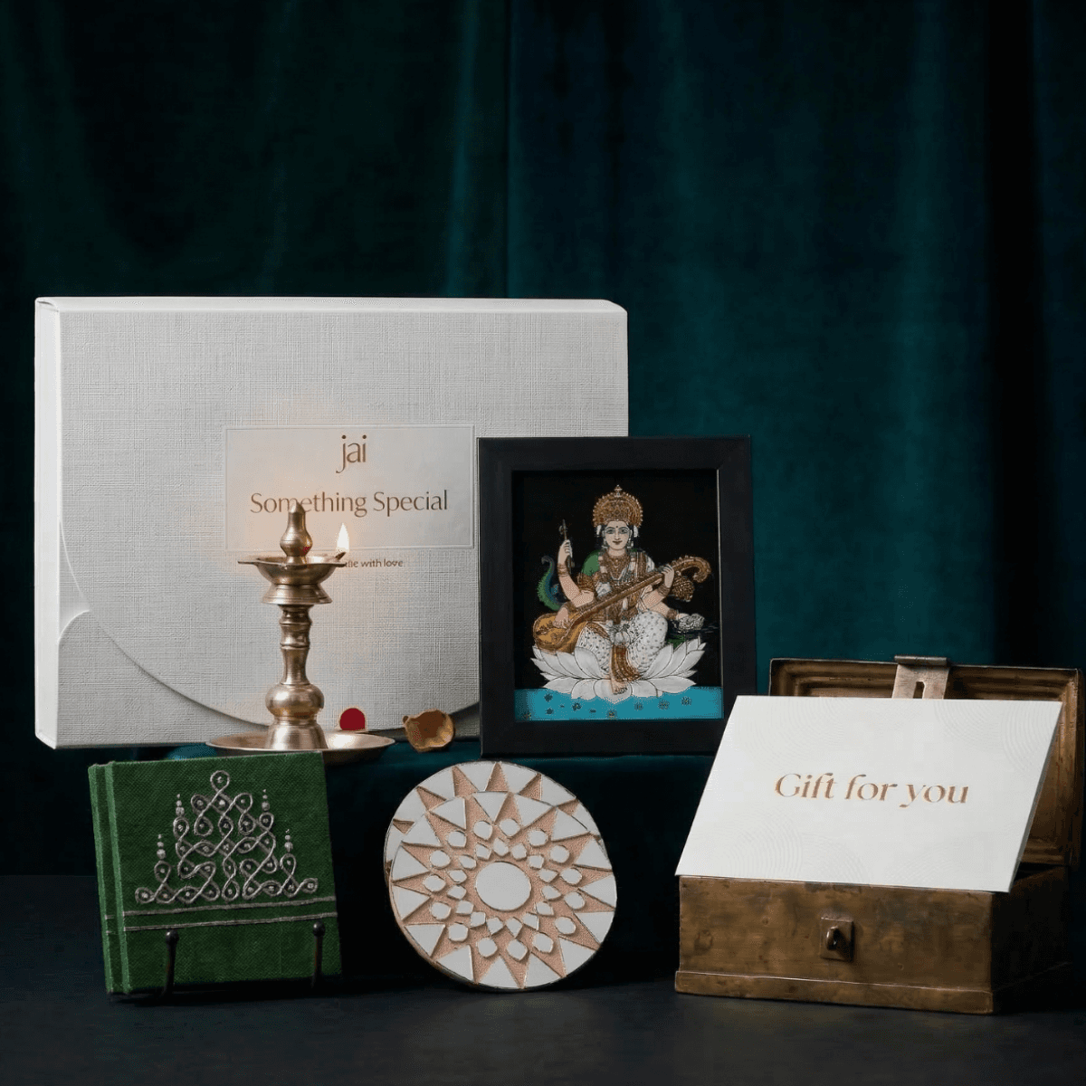 A premium white gift hamper box set featuring a reverse glass painting of Goddess Saraswati, two green hand-painted Manai, two mirror mosaic coasters, and a gift card, elegantly presented.