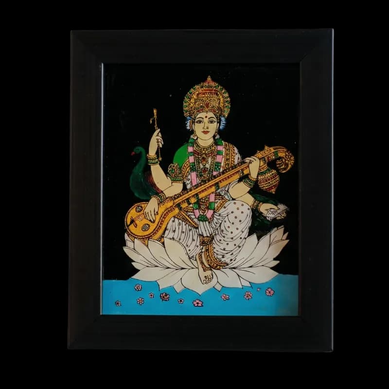 Handmade Goddess Saraswati reverse glass painting by Shree Jai Arts. The Hindu goddess of knowledge and music is beautifully depicted playing the Veena, seated gracefully on a white lotus with a peacock in the background, set against a dark backdrop within a premium black frame.